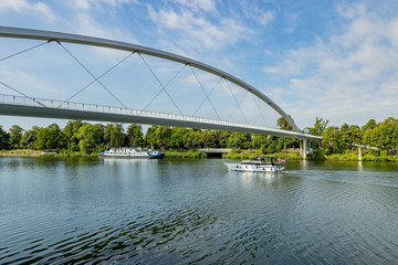 One boat sailing and another anchored on the bank of the Maas river surrounded by green trees and the High Bridge (Hoge Brug), sunny summer day with blue sky in Maastricht, South Limburg, Netherlands