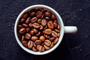 Top view of roasted whole coffee beans in white porcelain cup on gray textured background