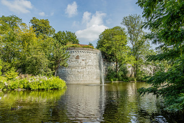 Fototapeta premium Pond with a small fountain surrounded by lush trees with the Rondeel (De Vijf Koppen) of the old city wall in the background, sunny day in the city park of Maastricht, South Limburg, Netherlands