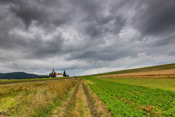 Dirt road leading to a small catholic church through agricultural field, gathering dramatic  storm clouds.