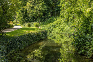 Jeker river next a path in the Maastricht city park, surrounded by trees and abundant green vegetation, with beautiful reflection in the calm waters on a sunny day in South Limburg, Netherlands
