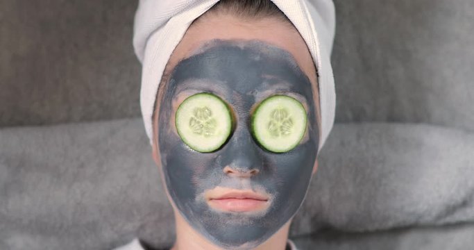 Young woman with grey mask on face is putting slices of cucumber on eyes. Her hairs wrapping in white towel. Spa beauty procedures at home for girl. Skincare, hydrating and moisturising concept.