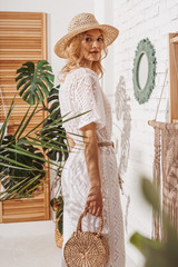 Young elegant fashionable lady wearing summer white crochet jumpsuit, straw hat, holding wicker bag, posing at home, in stylish boho interior with green tropical plants, trendy decorations  © Victoria Fox