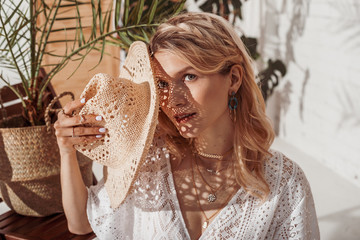 Summer fashion conception: elegant  woman enjoying sunlight, hiding her face with straw hat, wearing trendy earrings, necklace with many chains, posing in stylish boho interior. Without skin retouch