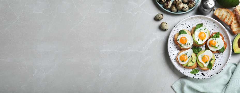 Flat lay composition of tasty bruschettas with quail eggs and avocado on light grey marble table, space for text. Banner design