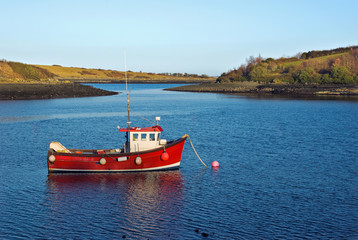 A little red fishing boat lies peacefully at anchor in a sheltered bay on a calm early Spring evening.