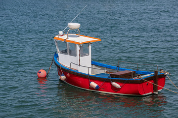 Obraz premium A little red fishing boat lies peacefully at anchor in a sheltered harbor on a warm Summer day.