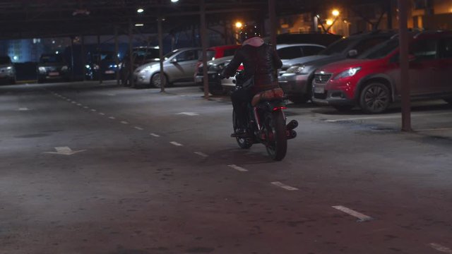 Slow motion rear view of biker in helmet and leather jacket hop on bike, start engine and driving away. Motorcyclist at parking garage riding motorbike at night, passing by parked cars