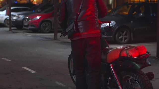 Slow motion of male biker, motorcyclist get on a bike at night. Rear view of motorcyclist in helmet and leather jacket going for a ride on motorbike, nighttime and red light on road