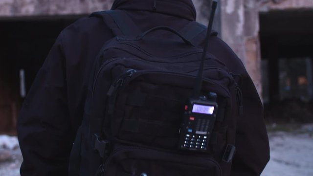 Slow-motion dolly shot from behind, security guard with walkie-talkie attached to backpack patrolling property, inspecting surroundings near restricted zone, bodyguard monitor abandoned building