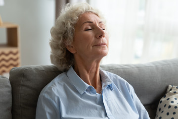 Head shot tranquil middle aged senior female retiree relaxing on comfortable sofa, doing morning yoga breathing exercises, enjoying calm peaceful moment indoors, daydreaming or visualizing future.
