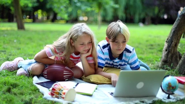 Stylish kids learning online outside in sunny park using laptop computer connecting online. 3G 5G internet connect. Boy and girl looking at notebook monitor resting on green grass. Technology.