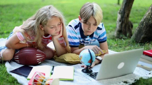 Traveling stylish kids learning geography online looking at Earth globe in green park using notebook. 5G internet connect. Boy and girl with notebook device resting on green grass. Laptop gadget.