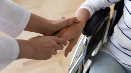 Fototapeta premium Close up medical social worker holding hand of disabled old retired woman sitting in invalid carriage, giving support or psychological help to handicapped ill patient indoors, rehabilitation concept.