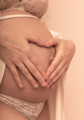 Closeup of pregnant woman holding her hands on her swollen belly shaping a heart, toned in pastel colors.