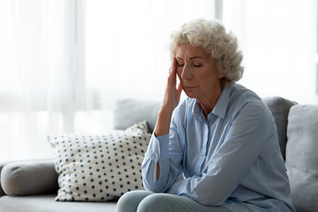 Exhausted mature old hoary woman sitting on sofa, suffering from headache indoors. Nervous middle aged elderly grandmother feeling doubtful about hard decision, health or psychological problems.