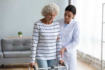 Focused middle aged hoary disabled woman using walking frame, making steps with help of professional physiotherapist. Young doctor helping older patient with body injury, rehabilitation process.