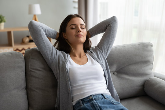 Young beautiful peaceful brunette woman leaning on comfortable sofa, stretching back with folded hands behind head, daydreaming with closed eyes, enjoying calm carefree moment alone in living room.
