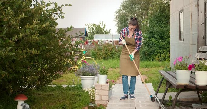 Woman Gardener In An Apron And Gloves Sweeps The Path With A Broom In Her Garden. A Young Girl Takes Care Of Her Dacha Plot. Working In Her Garden Near House.