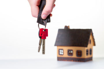 The hand holds the keys in the foreground. In the background, out of focus is a house against a light background. Business concept.