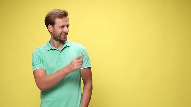 smiling young casual man holding hands in pockets, looking and pointing fingers to side, making thumbs up gesture and standing on pink background