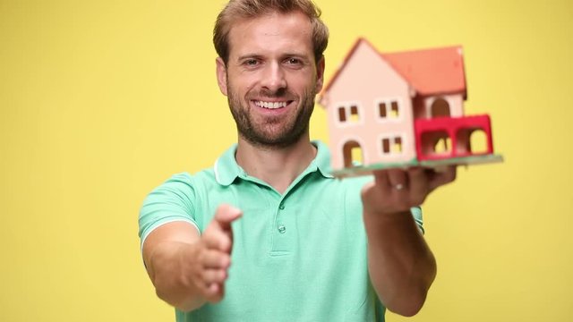 confident young casual man holding and presenting house, smiling, shaking hand and accepting deal on yellow background