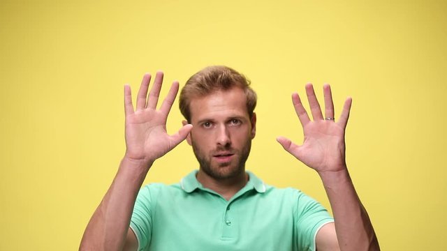 scared young casual man opening mouth in a scary manner, holding hands in the air and surrendering on yellow background