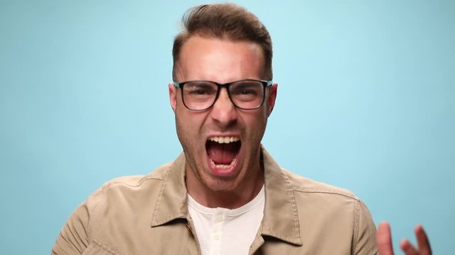 young sexy casual man leaning back then screaming out loud, touching his head and gesturing a mad person on blue background