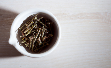 The screws lie in a white cup against a light background. With copy space.Top view.