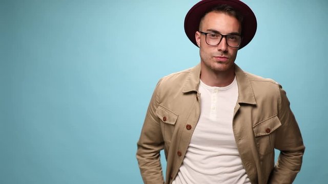 tough fashion model sticking his hands in his back pocket, wearing a burgundy hat, crossing his arms, fixing his eyeglasses and letting his arms fall loose on blue background