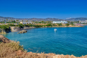A small lagoon with blue sea water on the coast of Paphos, Cyprus.  A small yacht is sailing in the creek.  In the distance you can see the hotel with the beach.