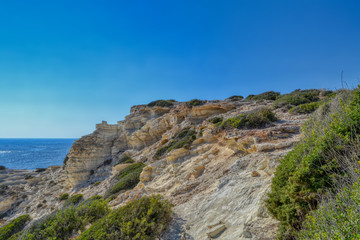 Fototapeta na wymiar Cliffs on the Mediterranean coast in the city of Paphos, Cyprus. Light stone blocks surrounded by small vegetation with the sea in the background.