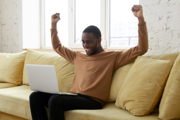 Happy black man excited getting good news notification at home. African american guy raising hands...