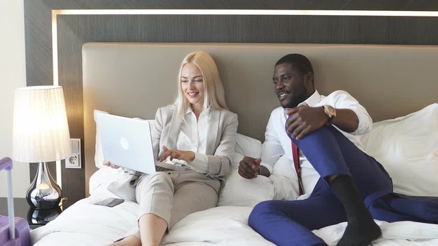 Beautiful multiethnic couple planning their trip, african guy and caucasian lady in formal wear lie on bed with laptop, talk.