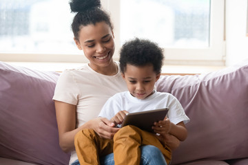 Once upon a time. African millennial mother sitting on sofa at living room holding toddler son on knees watching cartoon on tab pc screen, black nanny teaching little boy to read using education app