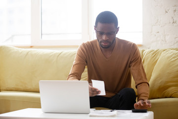 Unhappy african american man reviewing financial bill at home. Serious black male millennial reading invoice and calculating while planning budget or making finance operations from living room