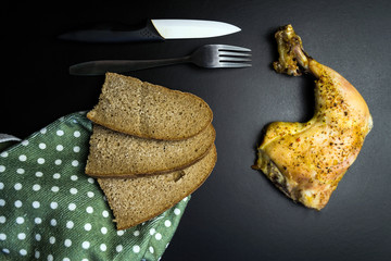 Baked chicken leg, rye bread, fork and knife. Top view on black
