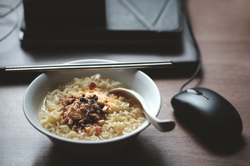instant noodles with seasoning garnish and Hot water in bowl on working space with laptop and mouse...