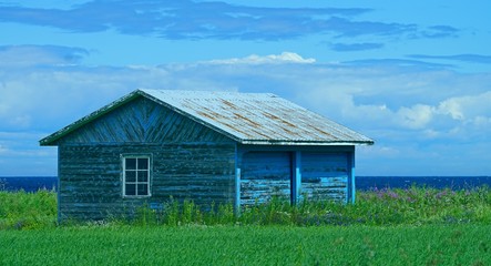 Very old barn by the sea  painted blue surrounded by green grass and a cloudy sky