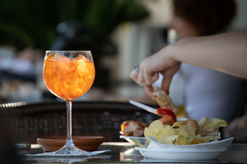 Close up of a Spritz, the typical northern italy drink or aperitif, made with sparkling white wine and orange spirit. Summer mood, outdoor table in a bar and a hand picking a snack from a plate.