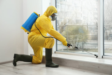 Pest control worker in protective suit spraying pesticide near window indoors