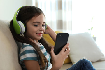 Cute little girl with headphones and smartphone listening to audiobook at home