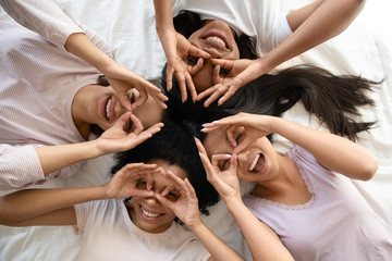 Group of cheerful diverse girls best friends lying in bed in pajamas making binoculars with fingers...