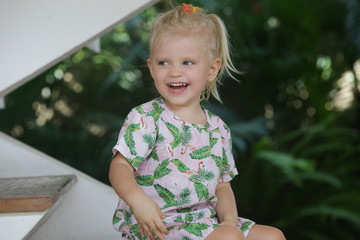 Adorable happy blonde toddler girl sitting at the wooden stairs at home in summer outfit.