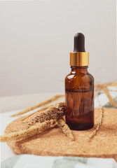 Brown cosmetic bottle with a pipette sits on a cork stand on a white table, with dried flowers nearby close up. Environmental materials. Warm colors.