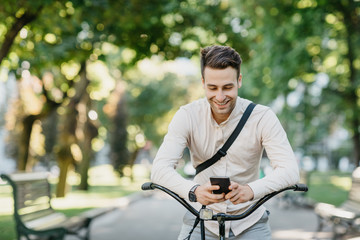 Social networks outdoors. Smiling attractive guy sits on bicycle in park and looks at smartphone