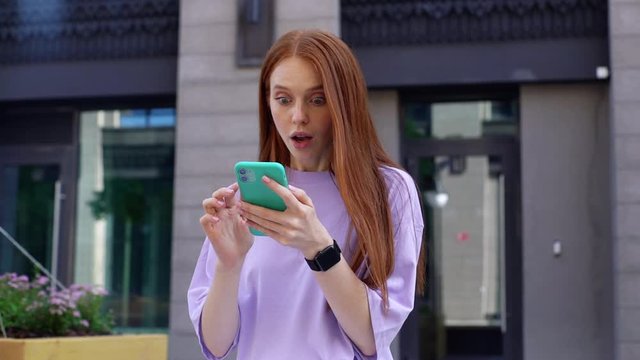 Surprised lady with red hair reading good news on phone outdoors background of city street. Portrait of excited beautiful woman checking phone content outside. Happy lady using smartphone in urban.
