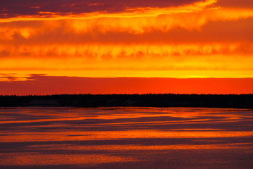 Fototapeta na wymiar A fiery sunset is reflected in the calm river water, overhanging clouds in the bright orange sky, light waves on the water surface.