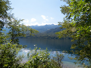 View of deep-blue lake with mountain range and blue sky in background and trees framing picture in foreground