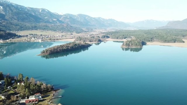 A drone flight above the Faaker See in Austria. There are high Alpine ranges around, reflecting in the crystal clear and calm surface of the lake. Small villages around the lake. Snow capped mountains